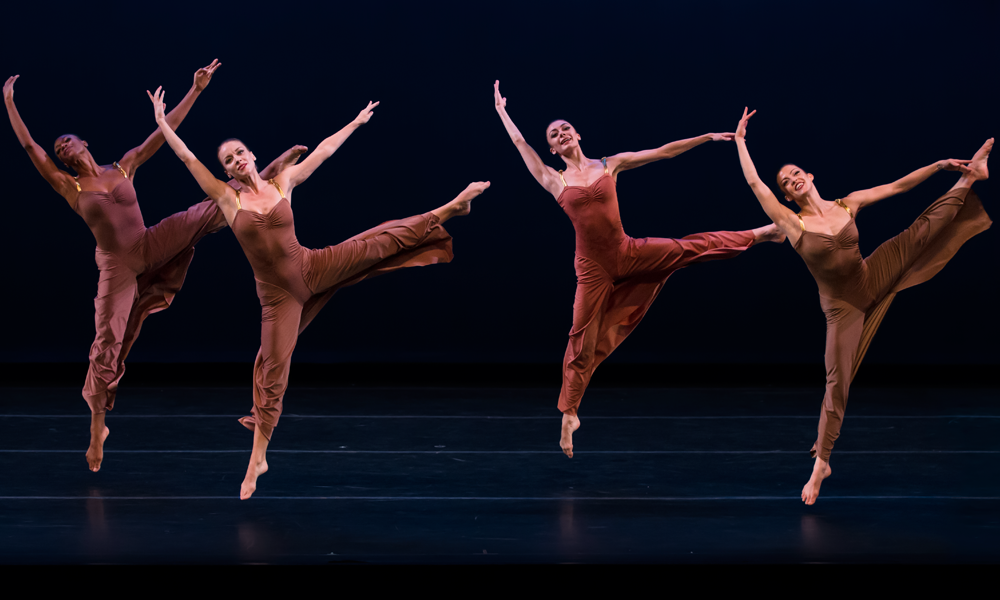 4 women dressed in rust colored gowns leap into the air their left legs extended their right legs pointing downwards. The expression here is of pure joy